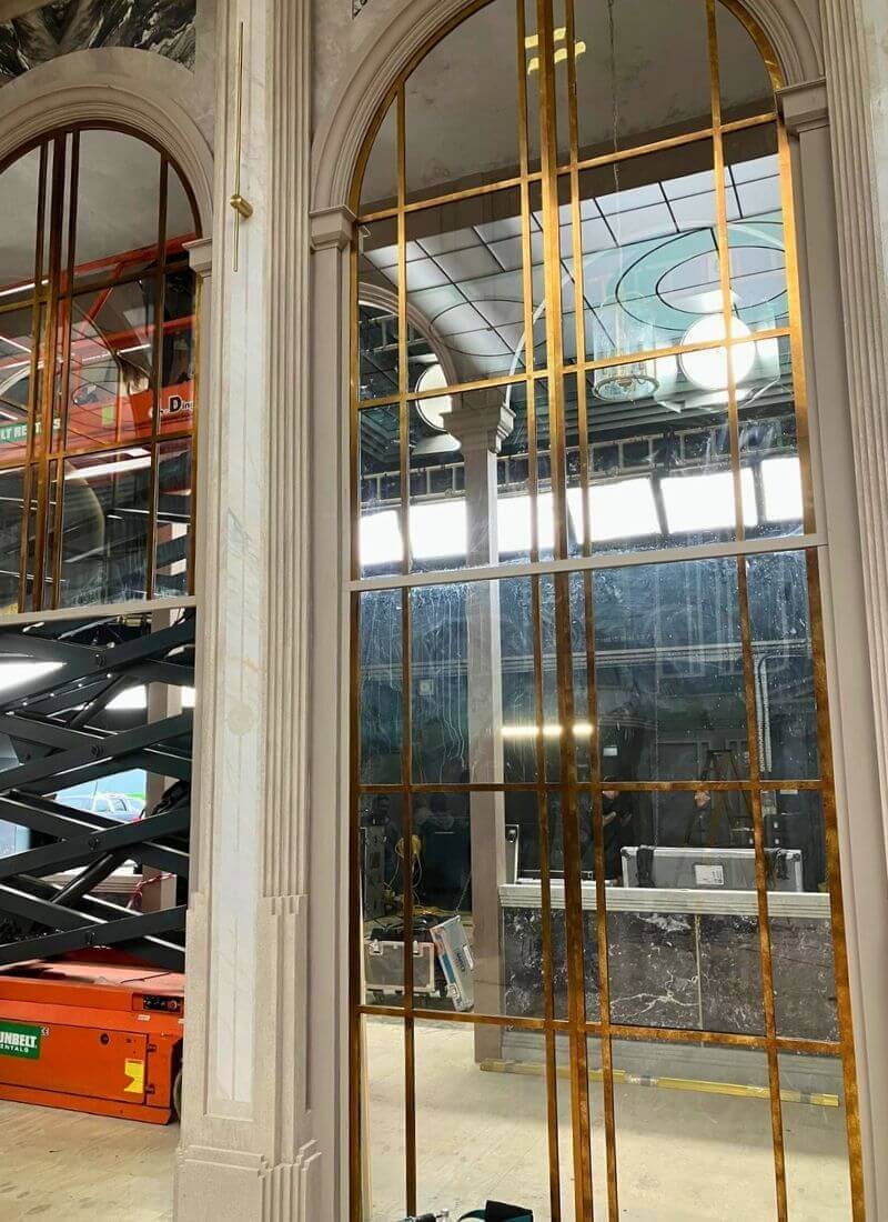 Man vs Baby - Window Metalwork During Construction Tall arched windows in mid-construction with gold-painted mullions and visible studio rigging behind.
